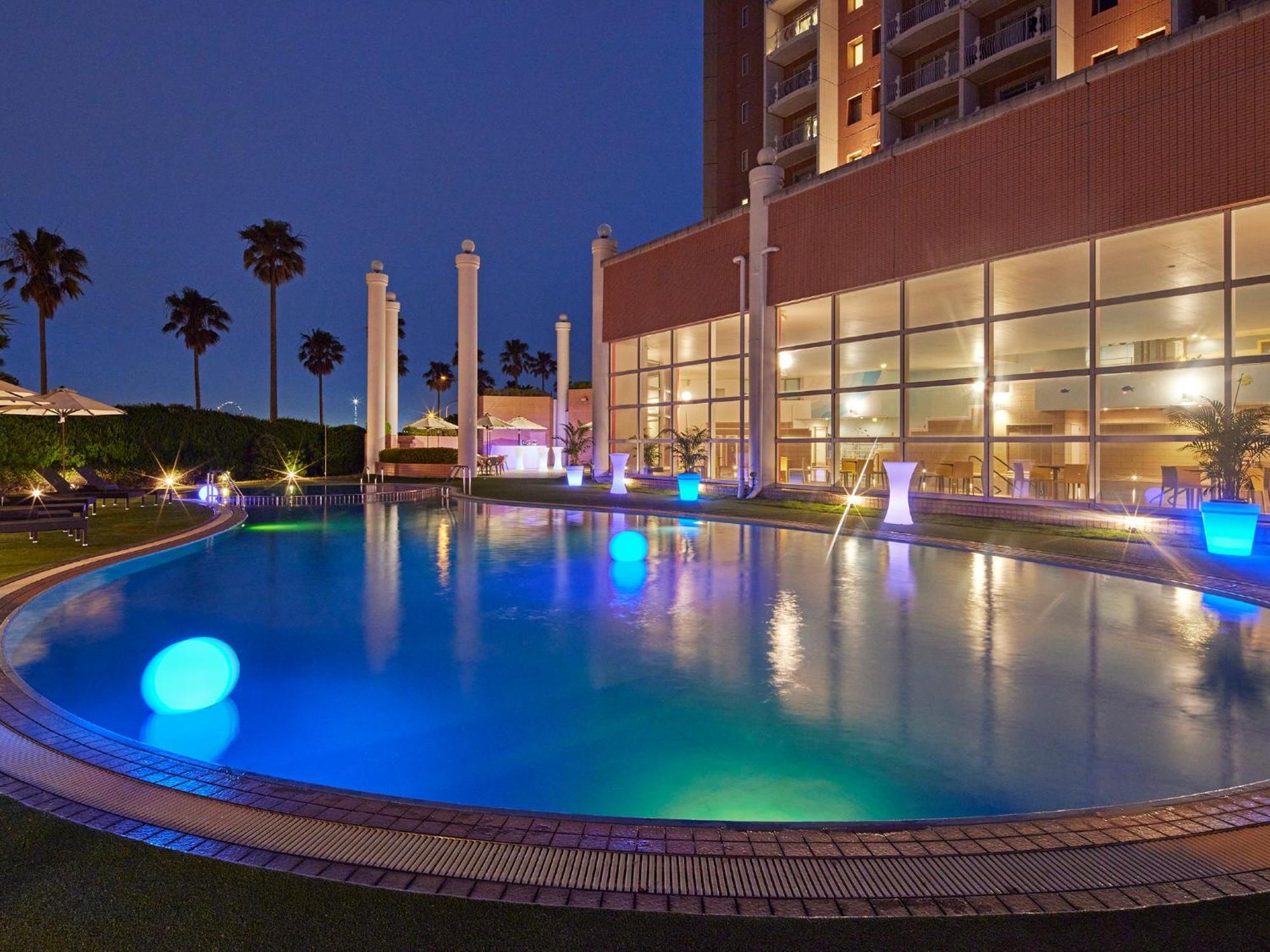 Grand Nikko Tokyo Bay Maihama Hotel Urayasu Eksteriør billede The photo shows a well-lit swimming pool at night, surrounded by a lush green area. The pool features glowing blue lights that create a serene and inviting atmosphere. Palm trees can be seen in the background, adding a tropical feel to the scene. Add