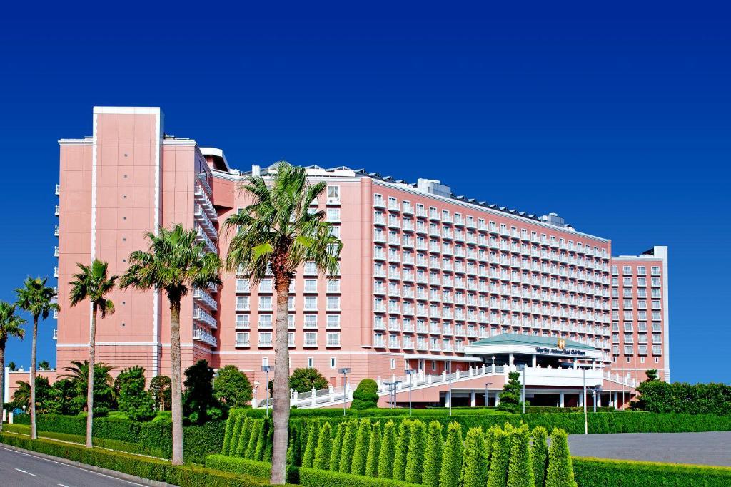 Grand Nikko Tokyo Bay Maihama Hotel Urayasu Eksteriør billede The image shows a large, multi-story hotel building featuring a light pink facade. The architecture appears modern, with numerous windows arranged in a grid pattern. In the foreground, there are neatly trimmed shrubs and palm trees, adding a tropical